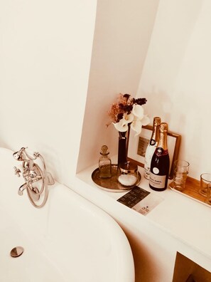 Un baño con bañera, tocador con espejo, una botella de champán y un jarrón con flores.