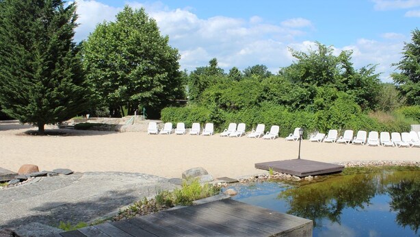 Een strand met witte ligbedden, een houten terras en een klein vijvertje.
