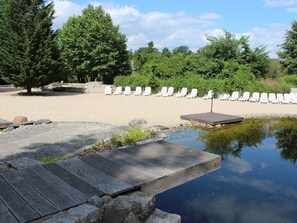 Une plage avec des chaises longues blanches, une terrasse en bois et un petit étang.