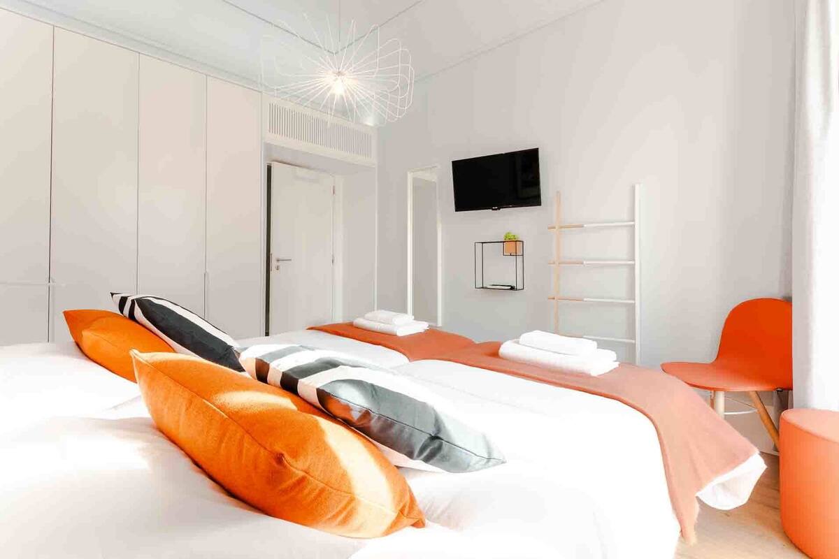 Ein modernes Hotelzimmer mit einem großen Bett, orangefarbenen und weißen Kissen, einem Flachbildfernseher, einem Schreibtisch und einem Stuhl.