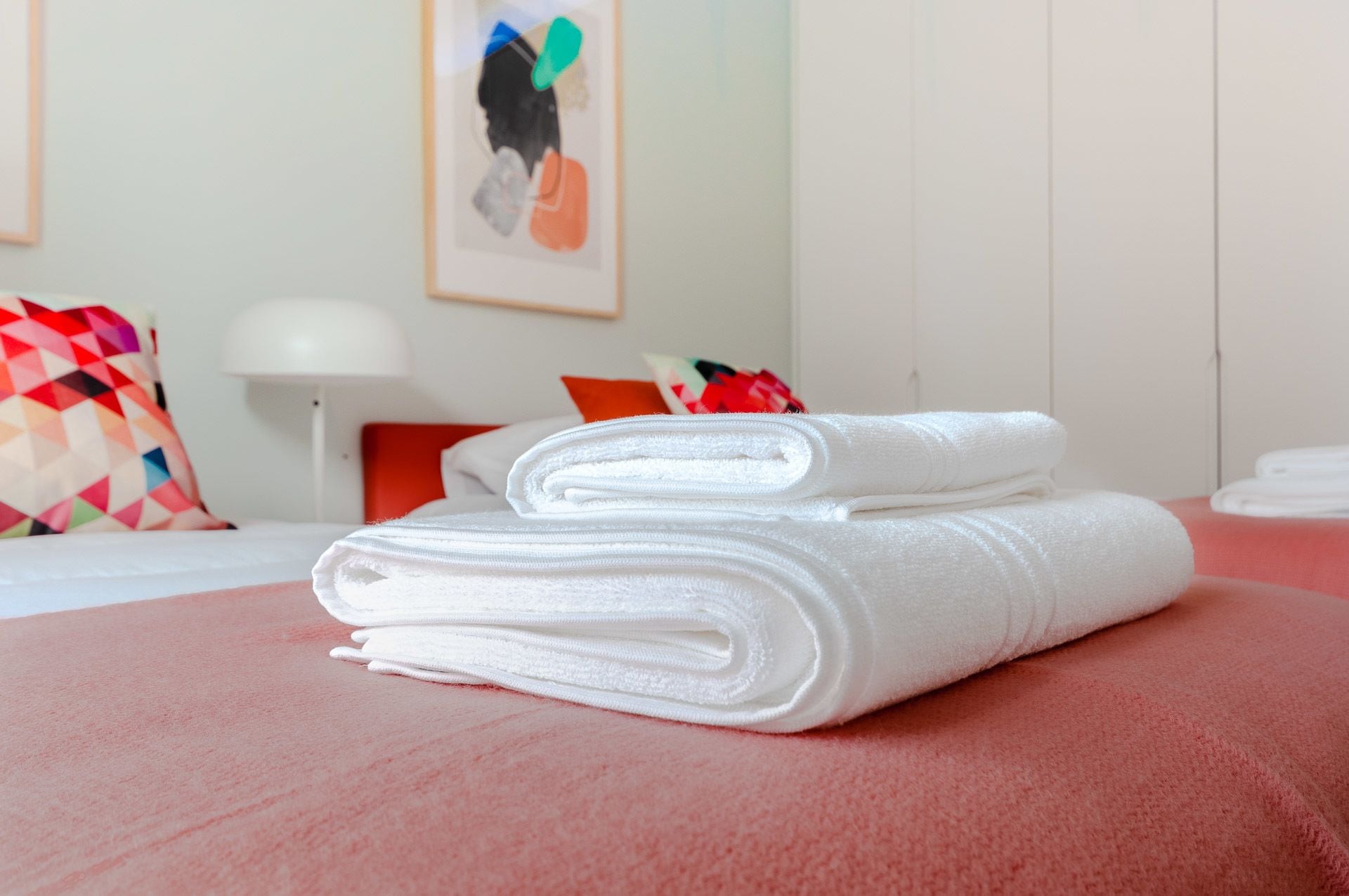 A neatly made bed with white towels, a red bedspread, and a colorful pillow.