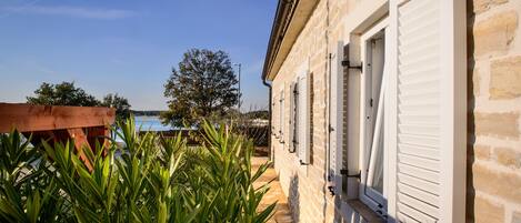 Un edificio de piedra con contraventanas blancas, una terraza de madera y vista al mar.