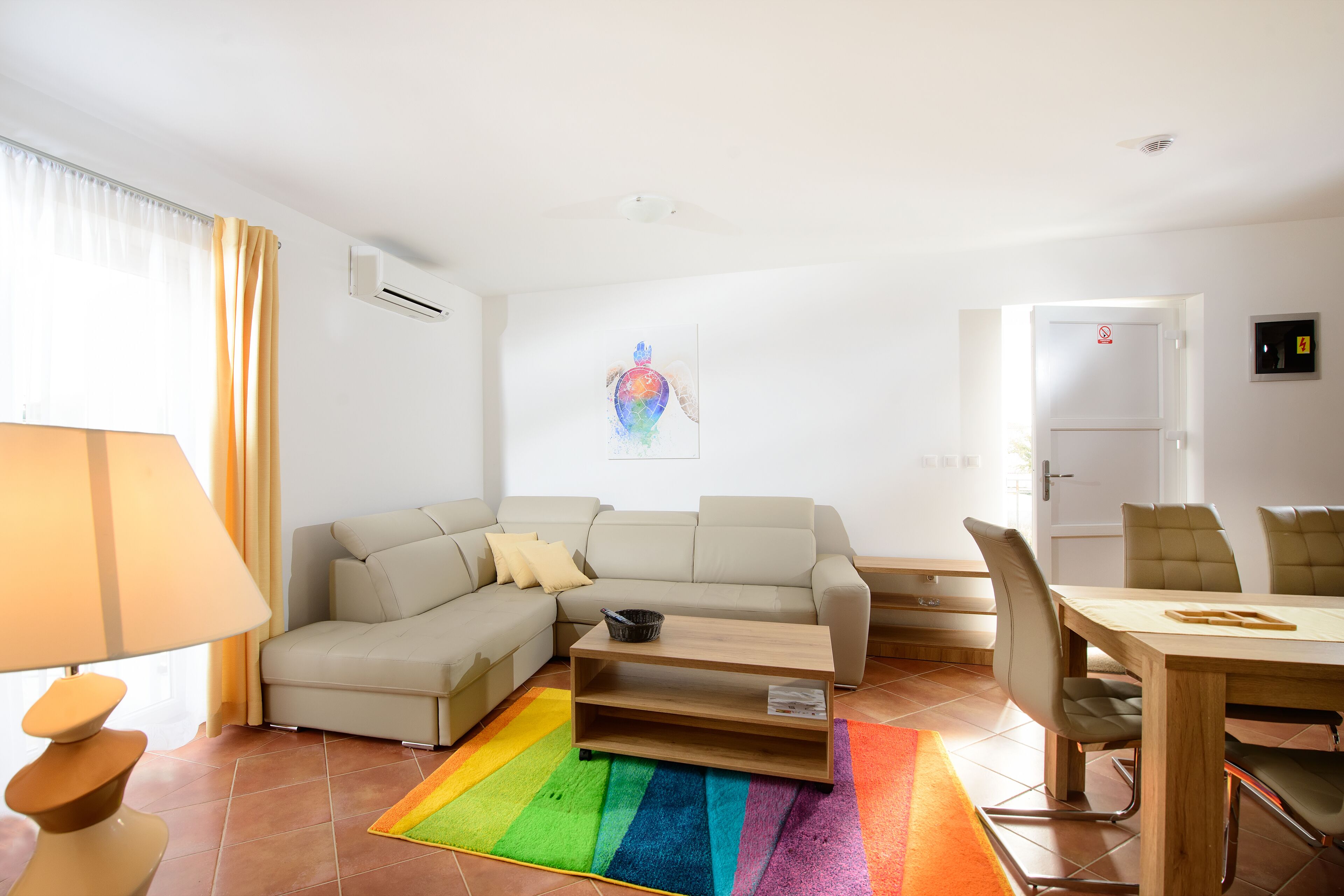 A living room with a sofa, a coffee table, a dining table with chairs, a colorful rug, and a wall-mounted air conditioner.