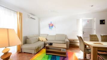 A living room with a sofa, a coffee table, a dining table with chairs, a colorful rug, and a wall-mounted air conditioner.