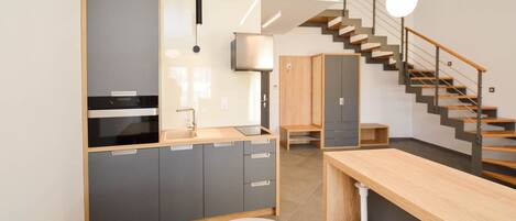A modern kitchen with grey cabinets, a wooden countertop, and a built-in oven. There is a staircase with a wooden railing leading to a landing.