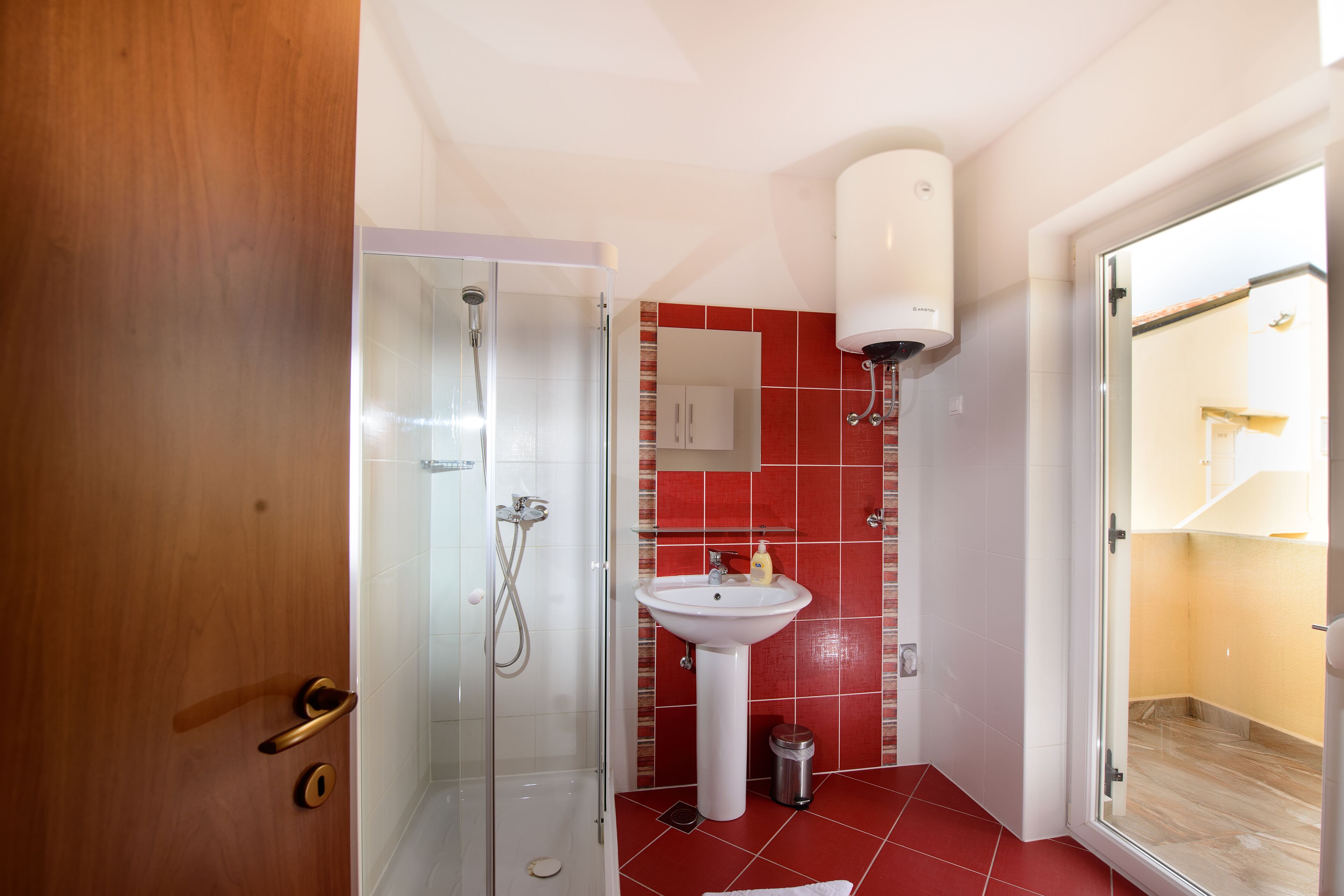 A bathroom with a glass shower enclosure, a pedestal sink, and a red-tiled floor.
