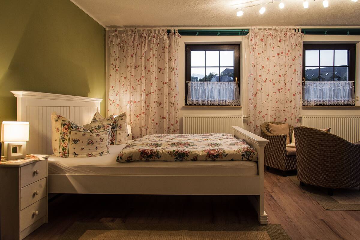 Ein Schlafzimmer mit einem Bett, einem Nachttisch, einem Sessel und zwei fensterhohen Vorhängen.