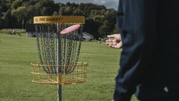 Una canasta de disc golf con cadenas y una red, ubicada en un campo de césped con un edificio al fondo.