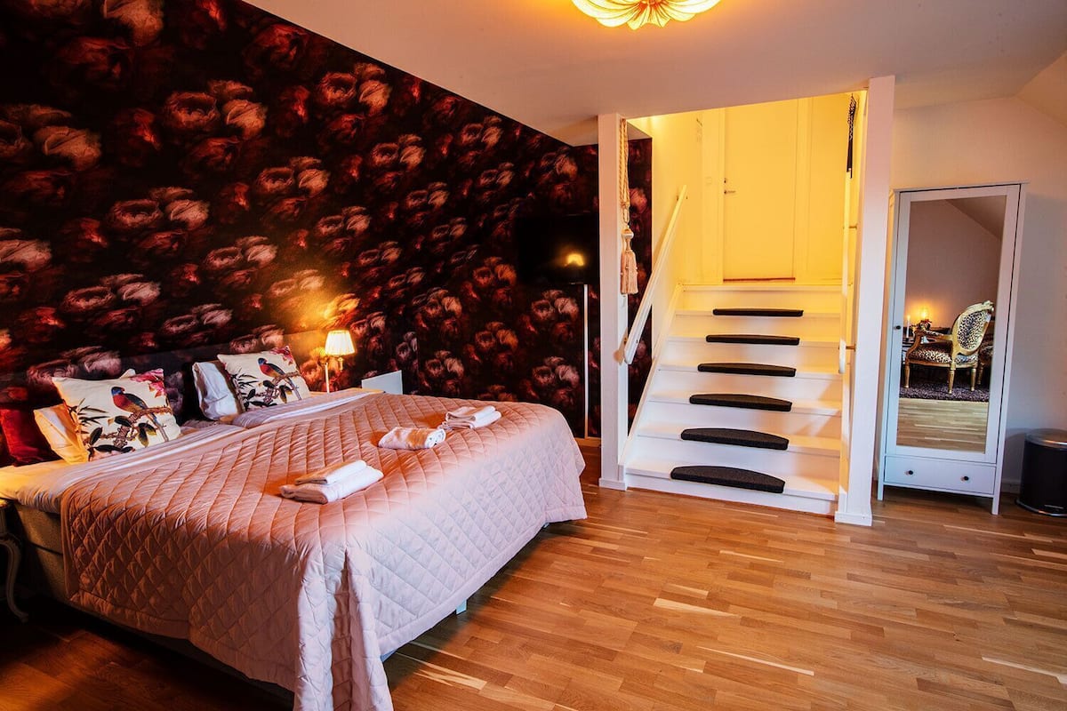 Um quarto com uma cama, uma mesa de cabeceira, um abajur, um espelho, uma escada e uma porta que dá para outro cômodo.