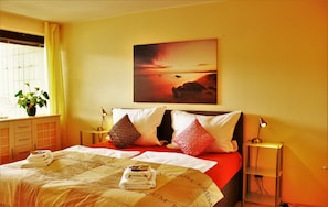 A hotel room with a bed, pillows, a nightstand, a lamp, a window with curtains, and a painting on the wall. - Weststadtblick (Heidelberg)