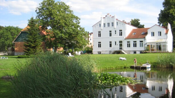 A large white building with a red-tiled roof, surrounded by greenery and a calm water body. - Gutshaus Gramkow (Hohenkirchen)