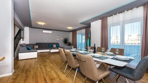 A modern dining area with a long table set for four, a grey sofa, and a view of the sea through large windows. - Amazing home in Sibenik (Sibenik)
