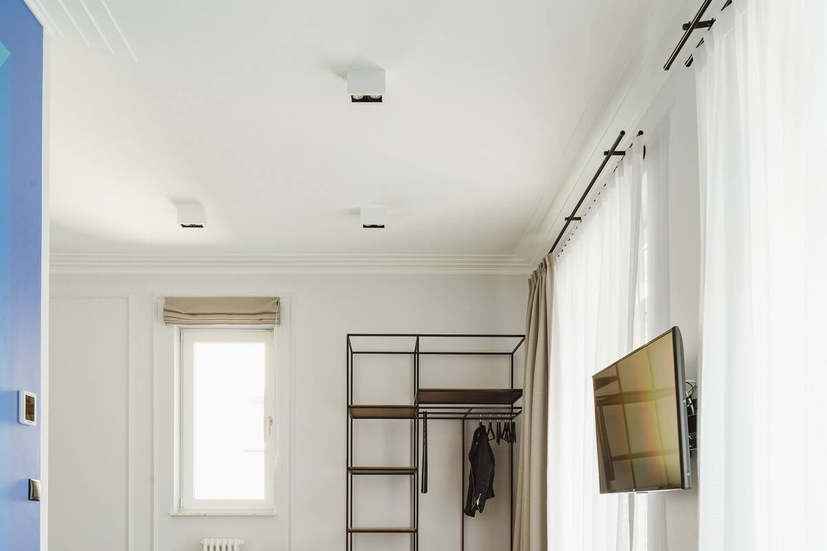 Una sala de estar moderna con un sofá azul, una mesa de centro de madera con un florero, una cama verde, un radiador, una ventana con cortinas, un televisor y un perchero.