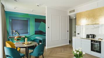 A modern room with teal and yellow chairs around a round table, a kitchen with white cabinets and a gold backsplash, and a vibrant abstract mural on the wall.