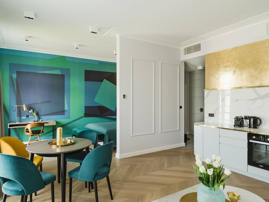 A modern room with teal and yellow chairs around a round table, a kitchen with white cabinets and a gold backsplash, and a vibrant abstract mural on the wall.
