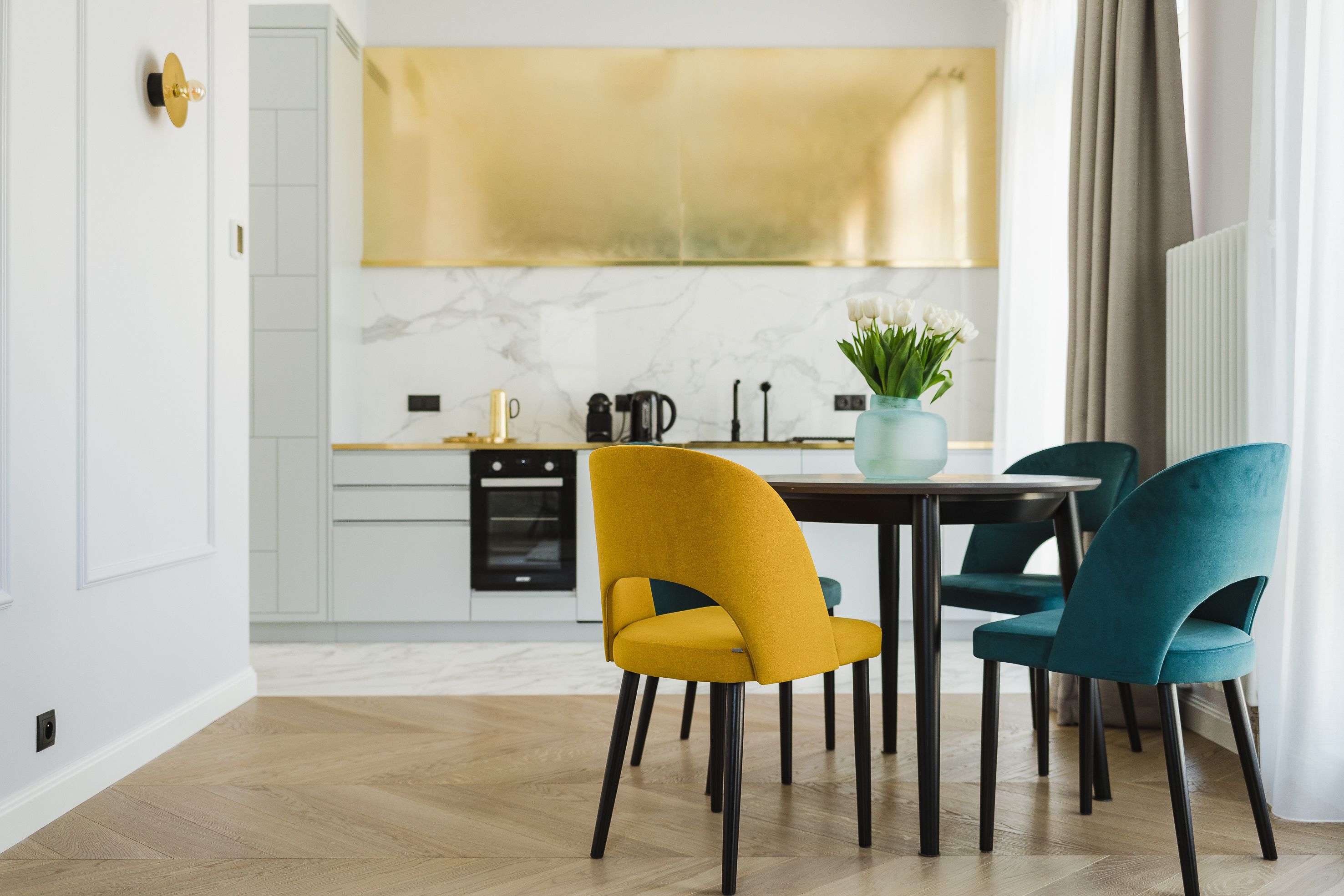 Une cuisine moderne avec des armoires blanches, un pare-feu en marbre et une hotte de couleur or. On y trouve deux chaises avec un revêtement jaune et turquoise, ainsi qu’un vase avec des fleurs blanches sur la table.