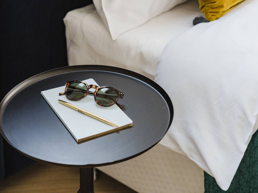 Una cama bien hecha con sábanas blancas y una almohada color mostaza, una pequeña mesa redonda con un cuaderno, bolígrafo y gafas de sol, y una cortina azul oscuro al fondo.