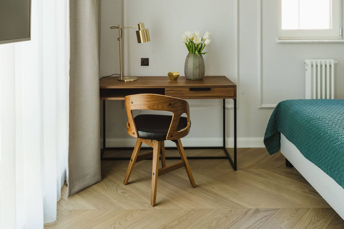 Un dormitorio con un escritorio de madera, una silla, una cama con manta verde, un radiador y una ventana con cortinas.