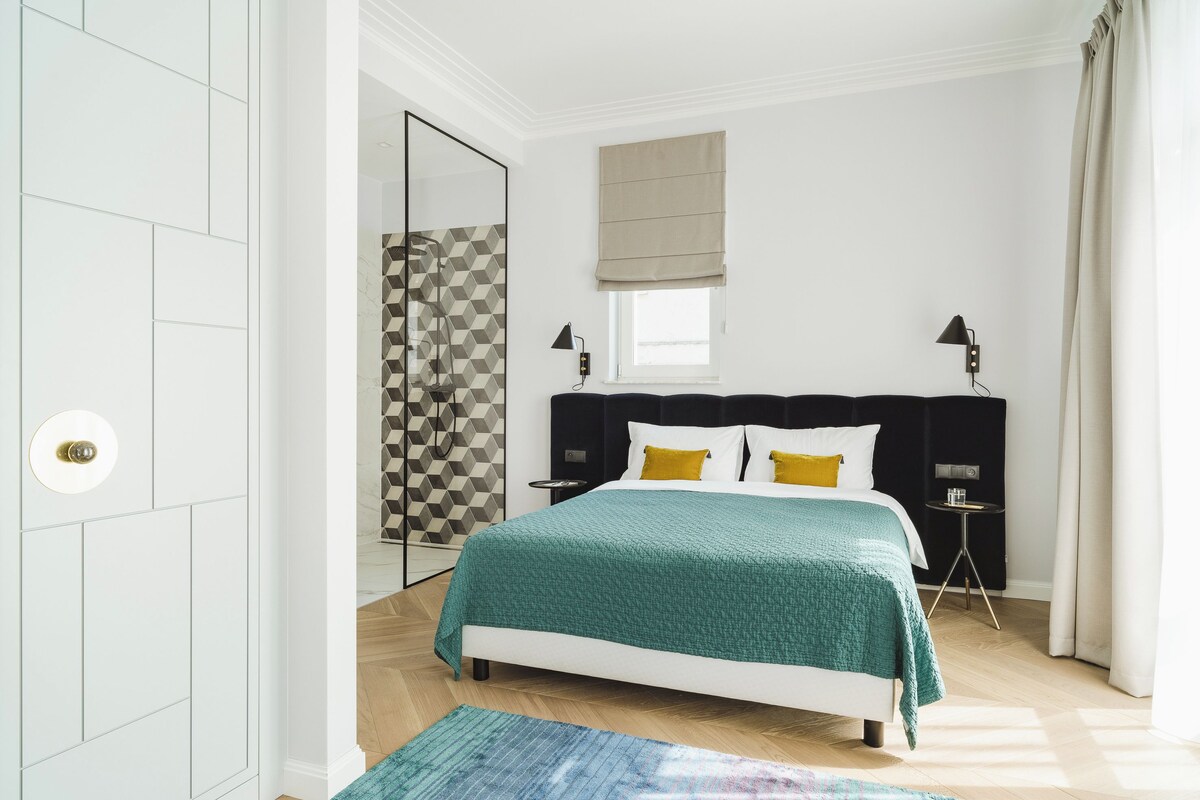 Un dormitorio con cama, mesitas de noche, una ventana con cortinas y un papel tapiz de diseño geométrico.