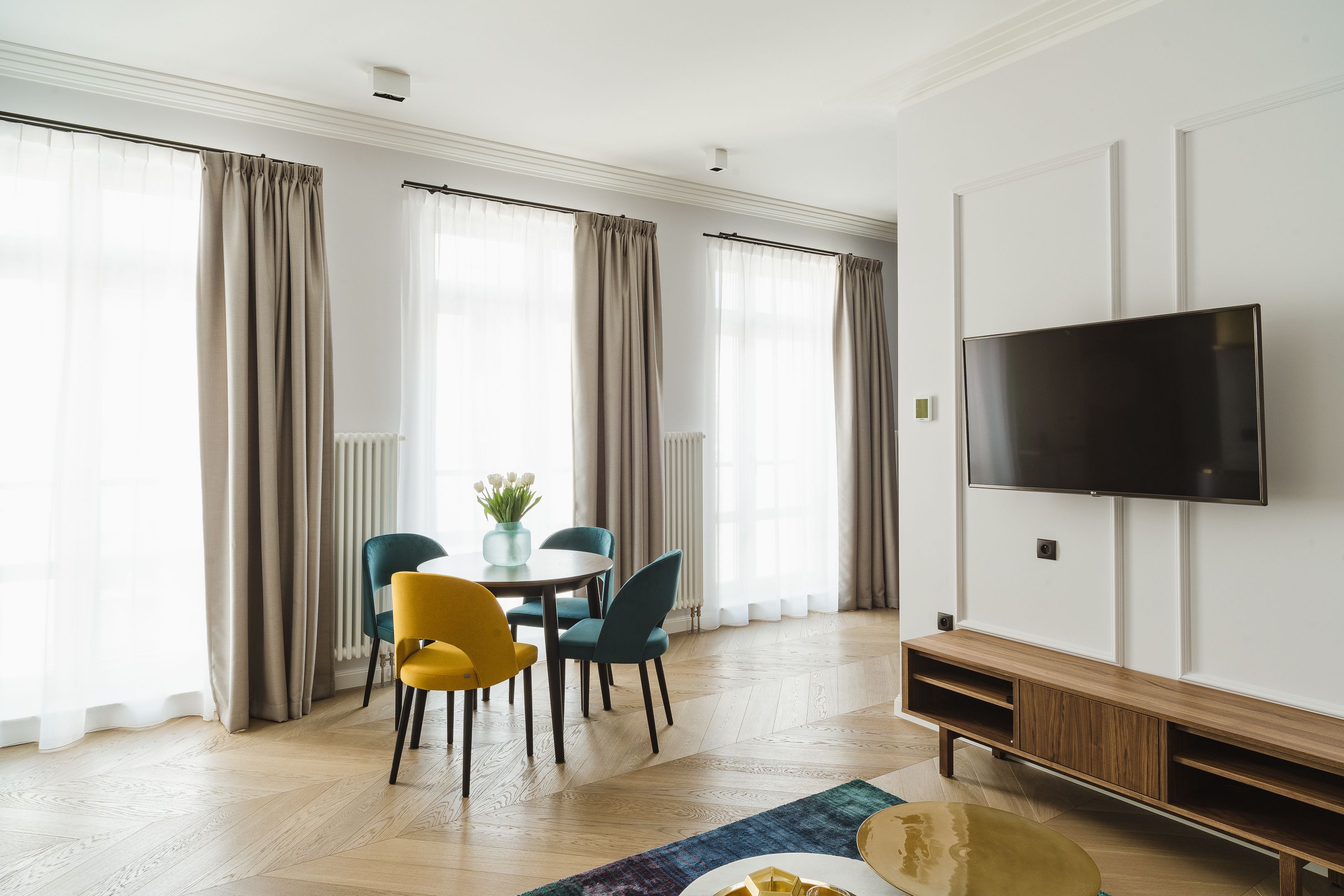 A modern living room with a dining area, a flat-screen TV mounted on the wall, and large windows with curtains.