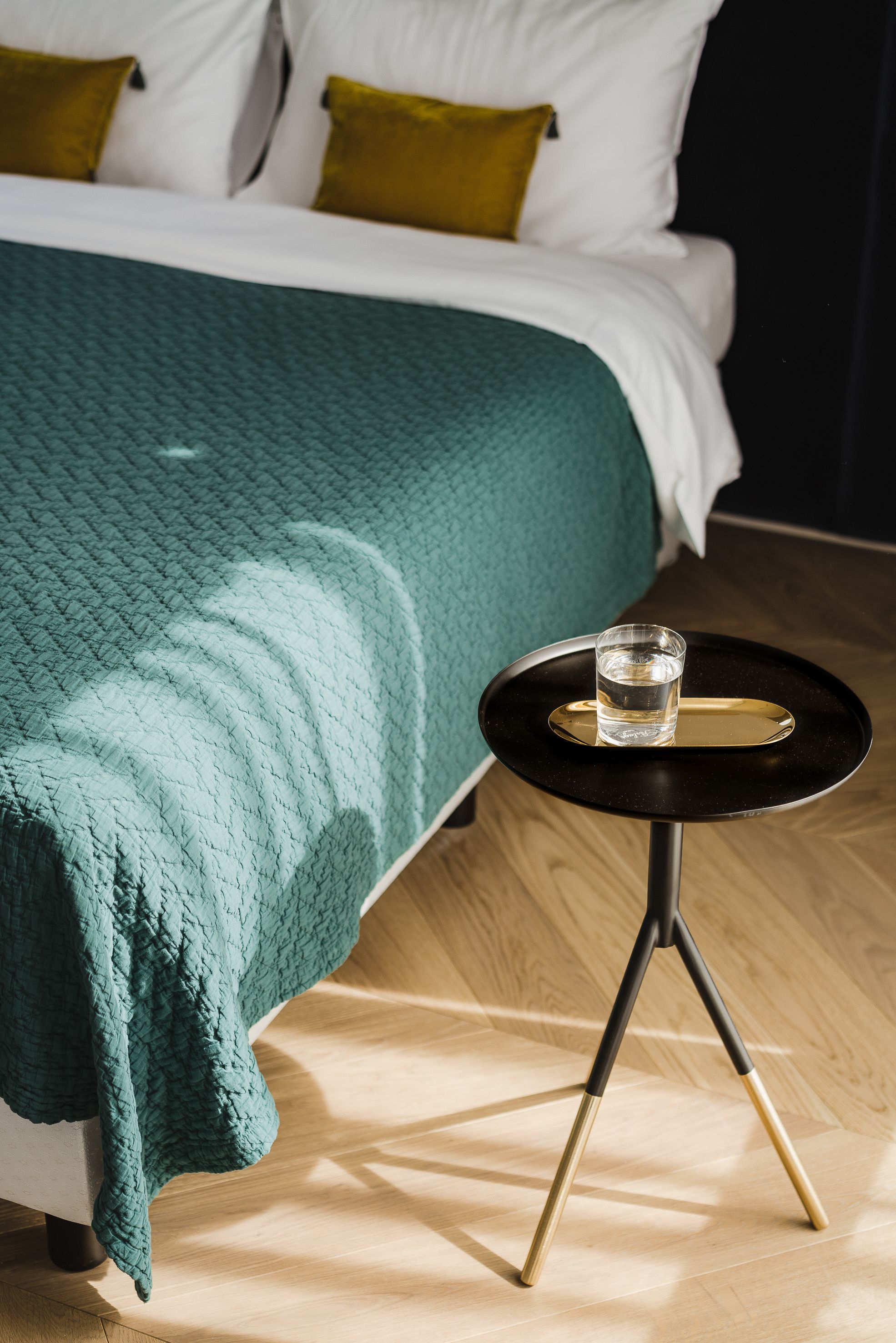 A neatly made bed with a teal blanket, a small round table with a glass of water, and a wooden floor.