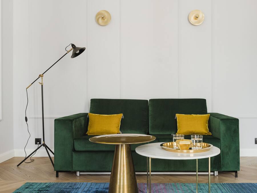 A green sofa with two yellow cushions, a round white table with a gold tray, and a floor lamp.