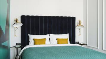 A neatly made bed with a teal blanket and two mustard pillows, flanked by two bedside tables with lamps, in a room with a dark headboard and white walls.