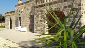 Un bâtiment en pierre doté d’une terrasse carrelée, de chaises blanches et d’une grande plante au premier plan.