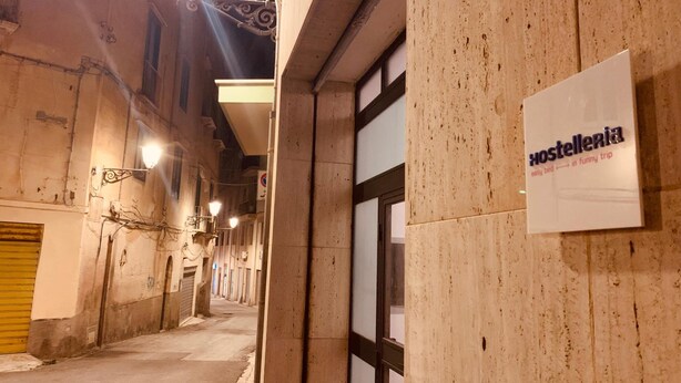 A narrow street with buildings on either side, a sign for 'Hostelleria' indicating a hostel, and a yellow shutter on the left. - Hostelleria (Trapani)