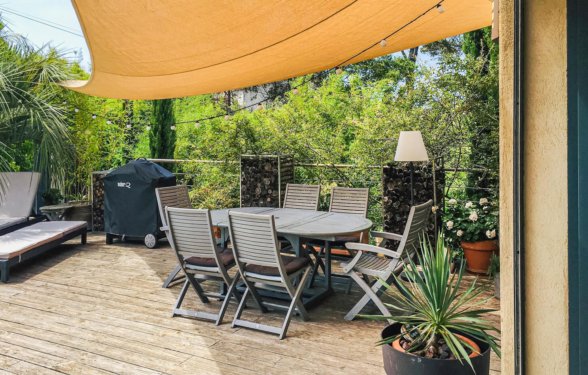An outdoor patio with a wooden deck, a round table with chairs, a barbecue grill, and potted plants.