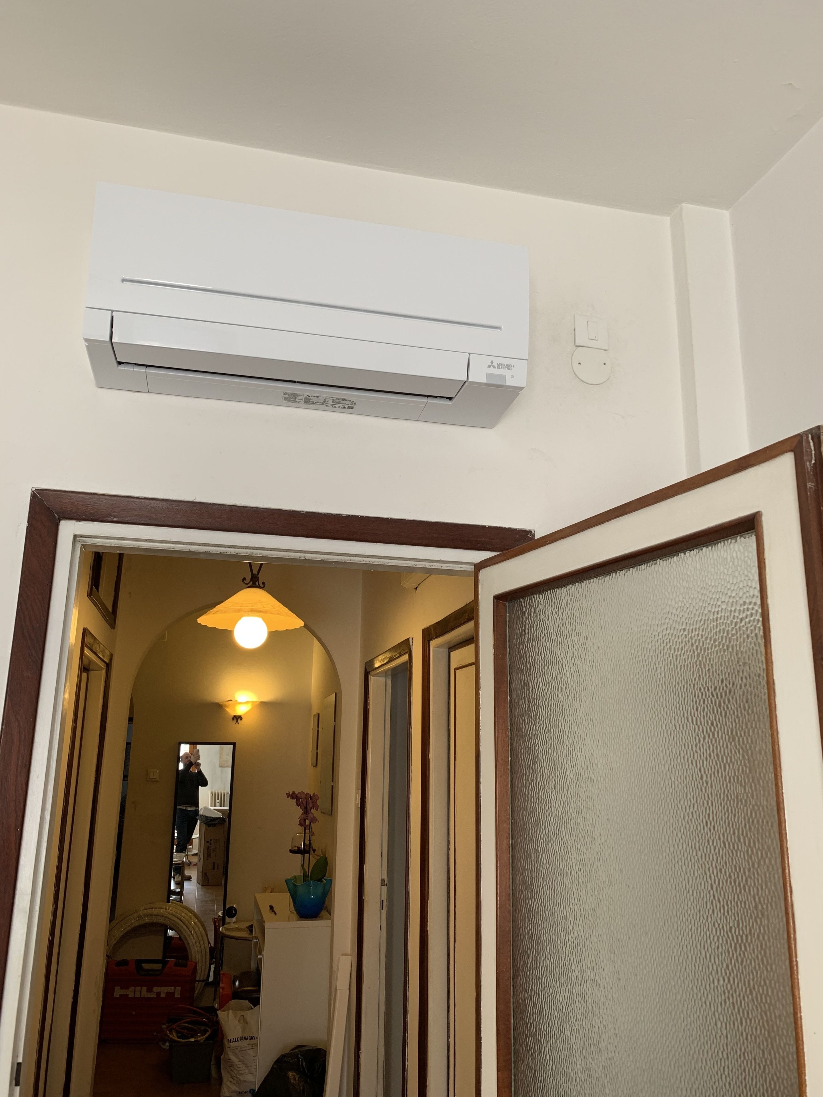 A hallway with a white air conditioning unit mounted on the wall, a wooden-framed door, and a glass panel on the right.