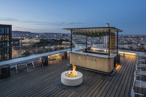 A rooftop bar with a fire pit, bar counter, and seating area overlooking a cityscape.