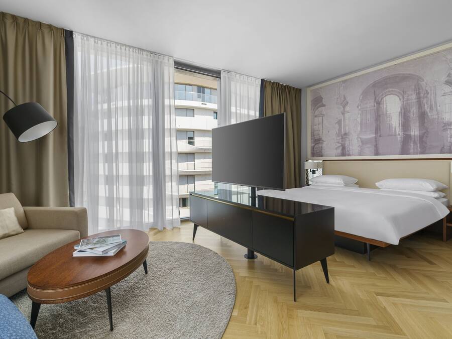 Habitación de hotel con cama, televisor, sofá, mesa de centro y un ventanal con cortinas.
