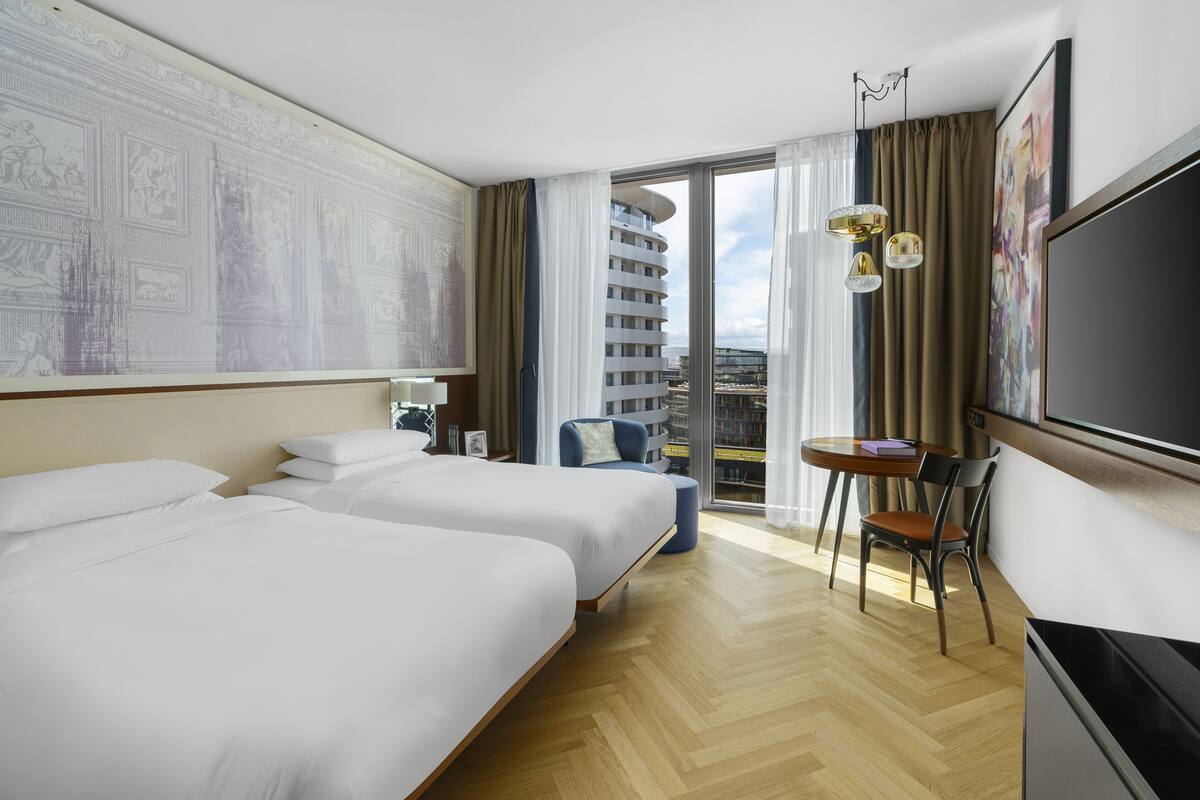 Habitación de hotel con dos camas, televisión, escritorio con silla, balcón con vista a un edificio y una obra de arte grande en la pared.