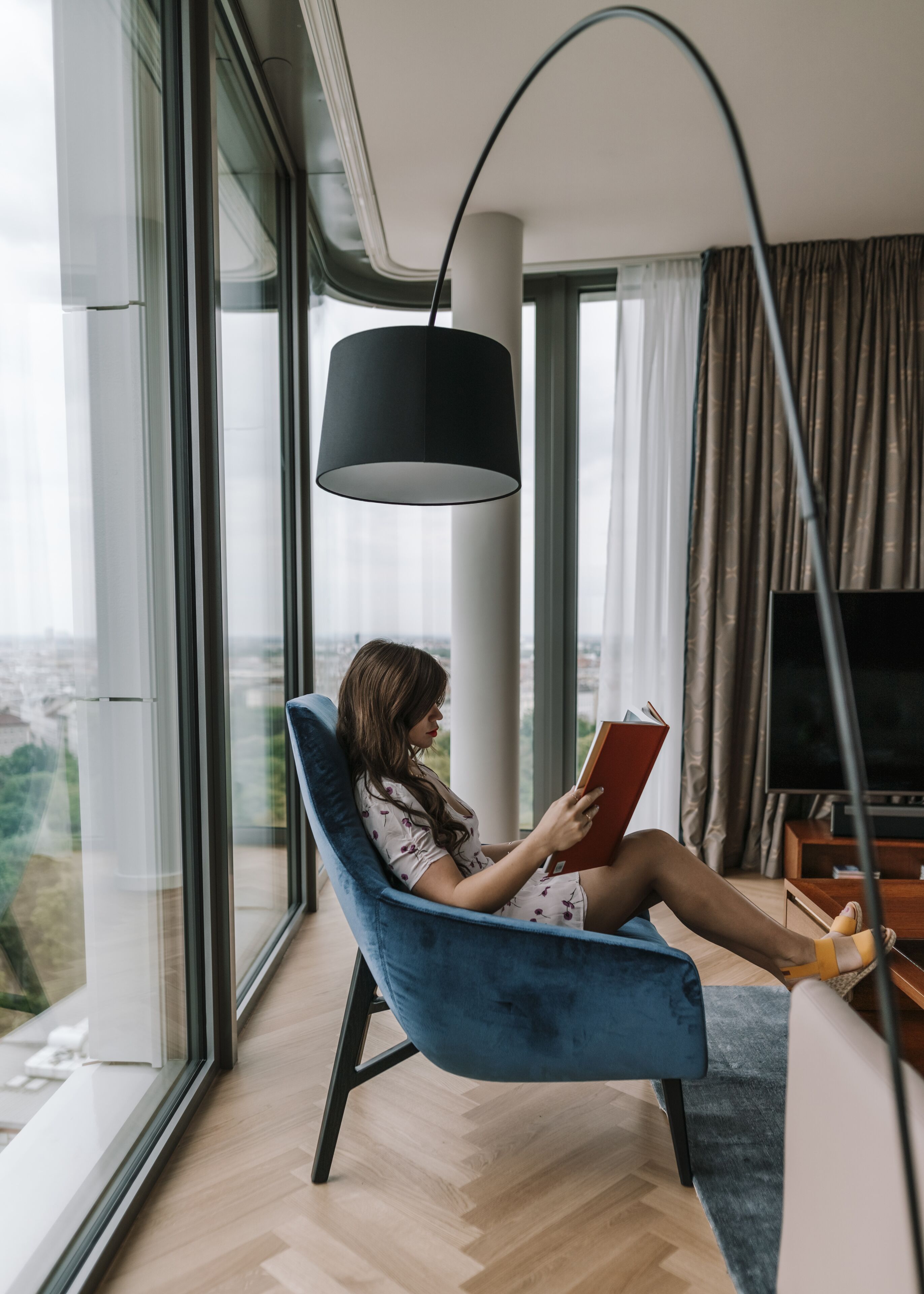 Una mujer sentada en una silla azul leyendo un libro en una habitación moderna con ventanas amplias y una lámpara de pie negra.