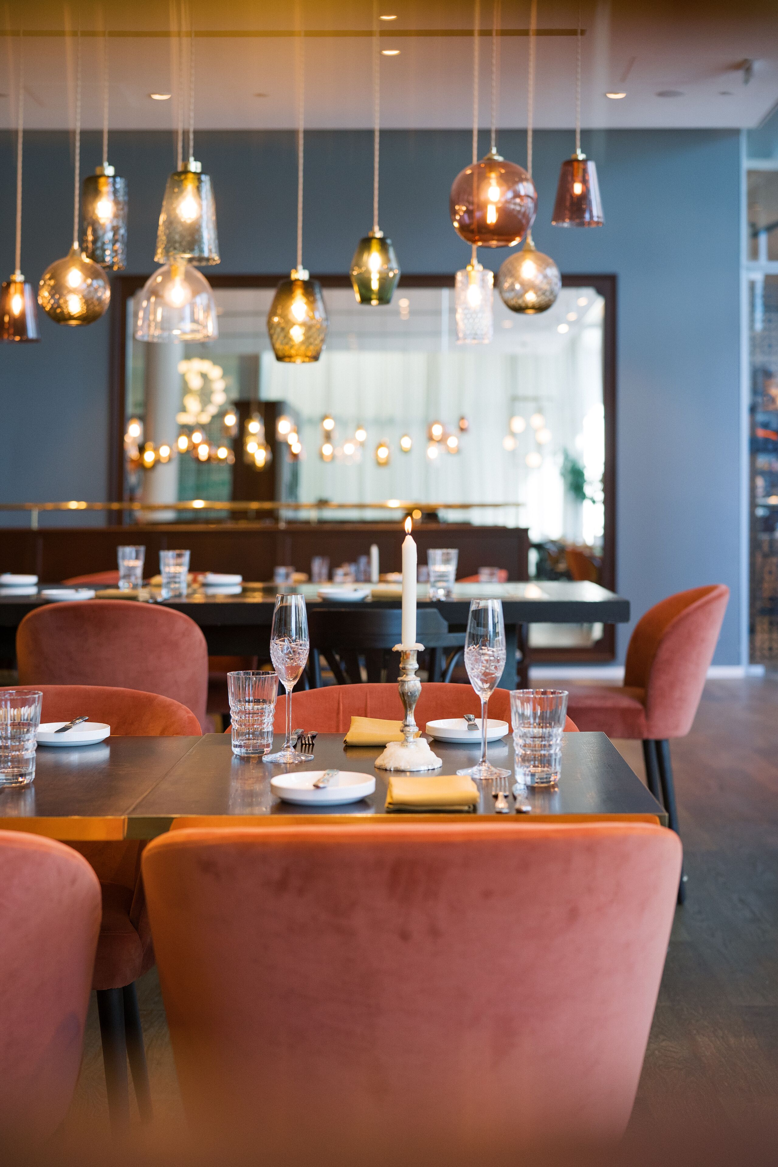 A dining area with a long table set for two, surrounded by plush chairs. Overhead, there are multiple hanging pendant lights.