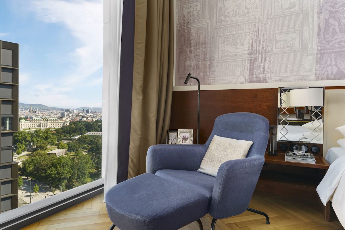 Habitación de hotel moderna con un ventanal grande que ofrece vistas a la ciudad, un sillón azul cómodo, una mesita de noche de madera y una cama con ropa de cama blanca.