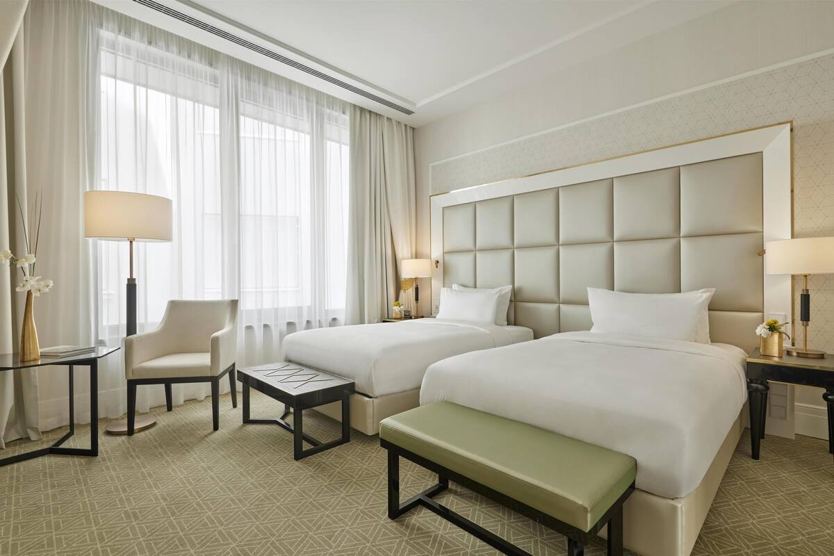 A hotel room with two beds, a bench, a chair, a small table, and a lamp.