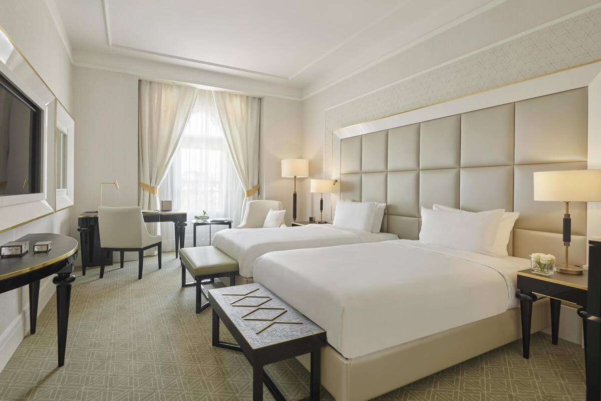 A hotel room with two beds, a desk, a chair, a small table, and a TV.