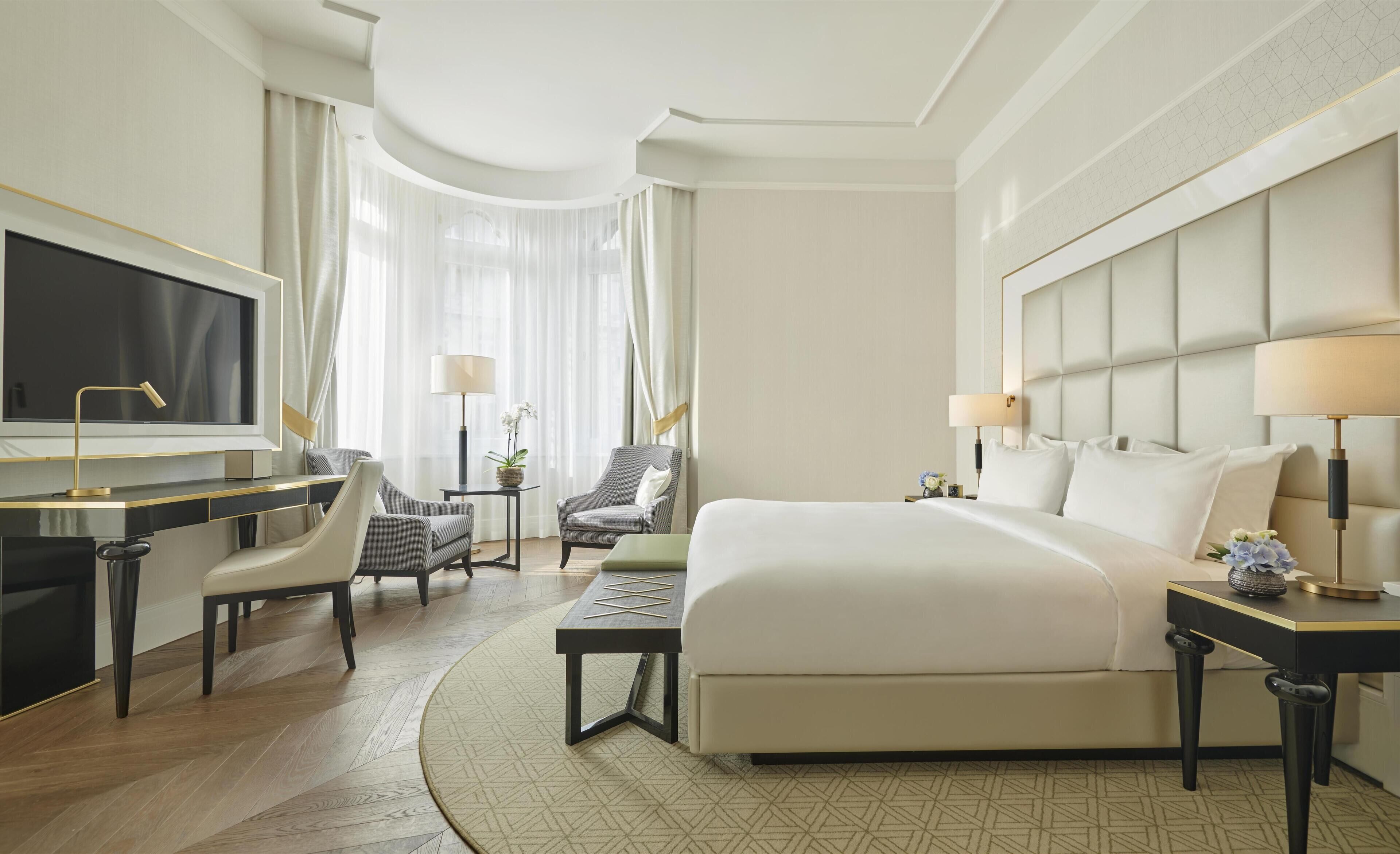 Un cuarto de hotel con una cama grande, un escritorio con televisor, dos sillones, una mesita pequeña y una mesita de noche con lámpara.