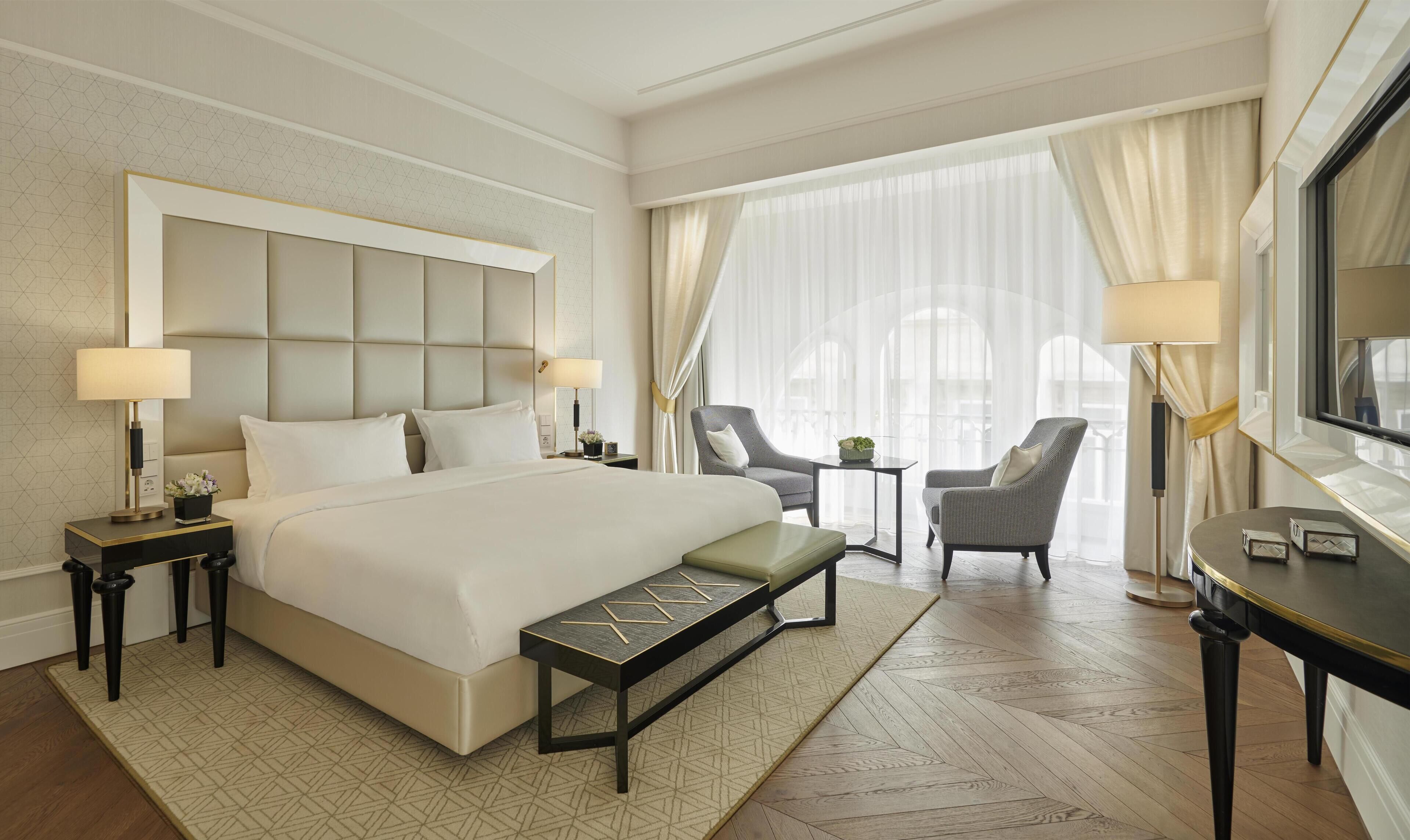 Habitación de hotel con una cama grande, dos mesitas de noche con lámparas, una silla, una pequeña mesa con una planta y un ventanal con cortinas.