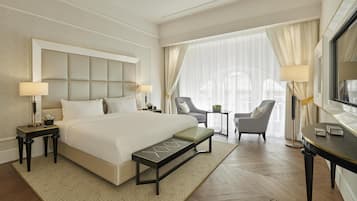 Habitación de hotel con una cama grande, dos mesitas de noche con lámparas, una silla, una pequeña mesa con una planta y un ventanal con cortinas.