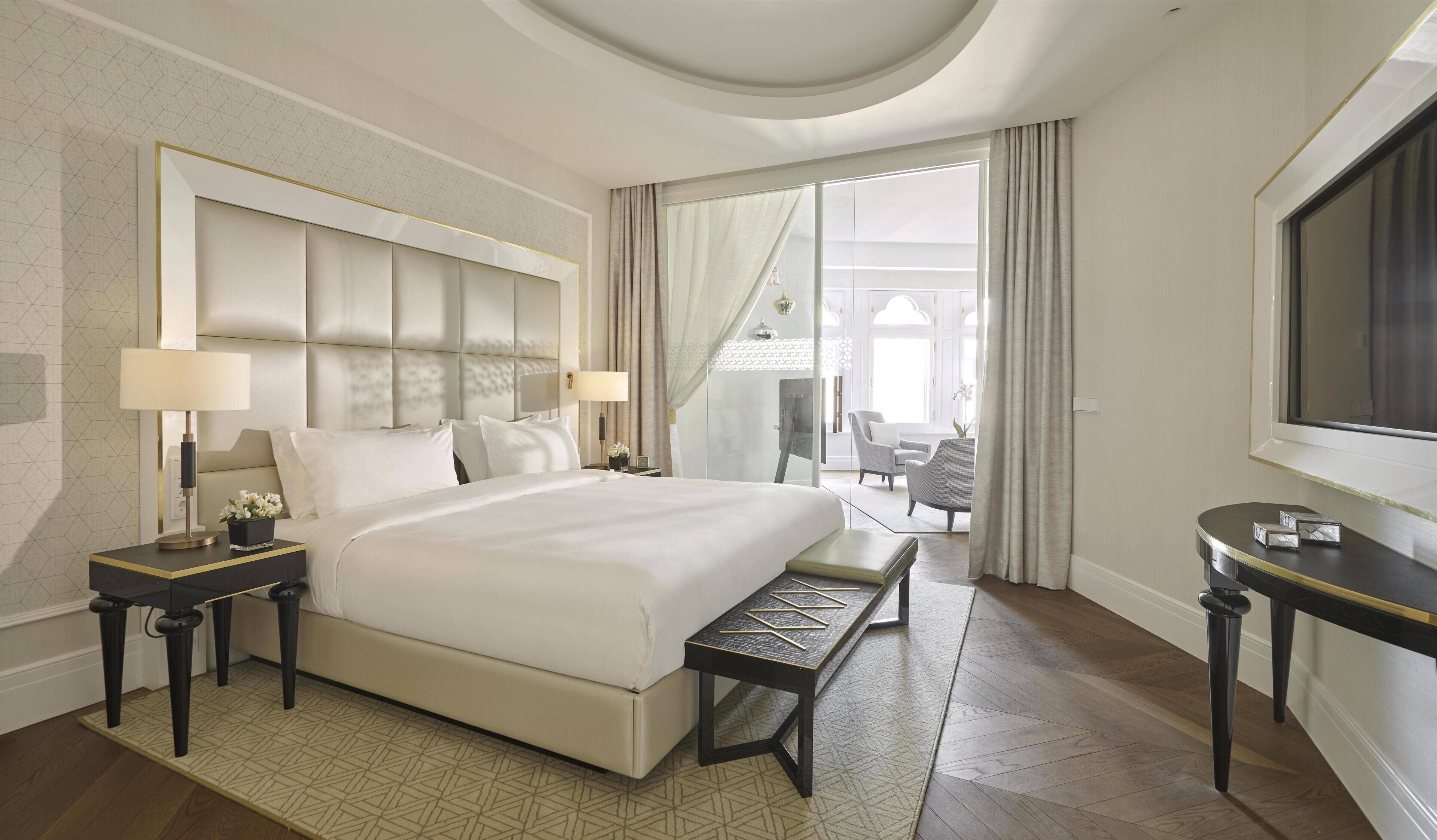 A modern hotel room with a large bed, bedside tables with lamps, a bench, a TV, and a view of the outdoors.