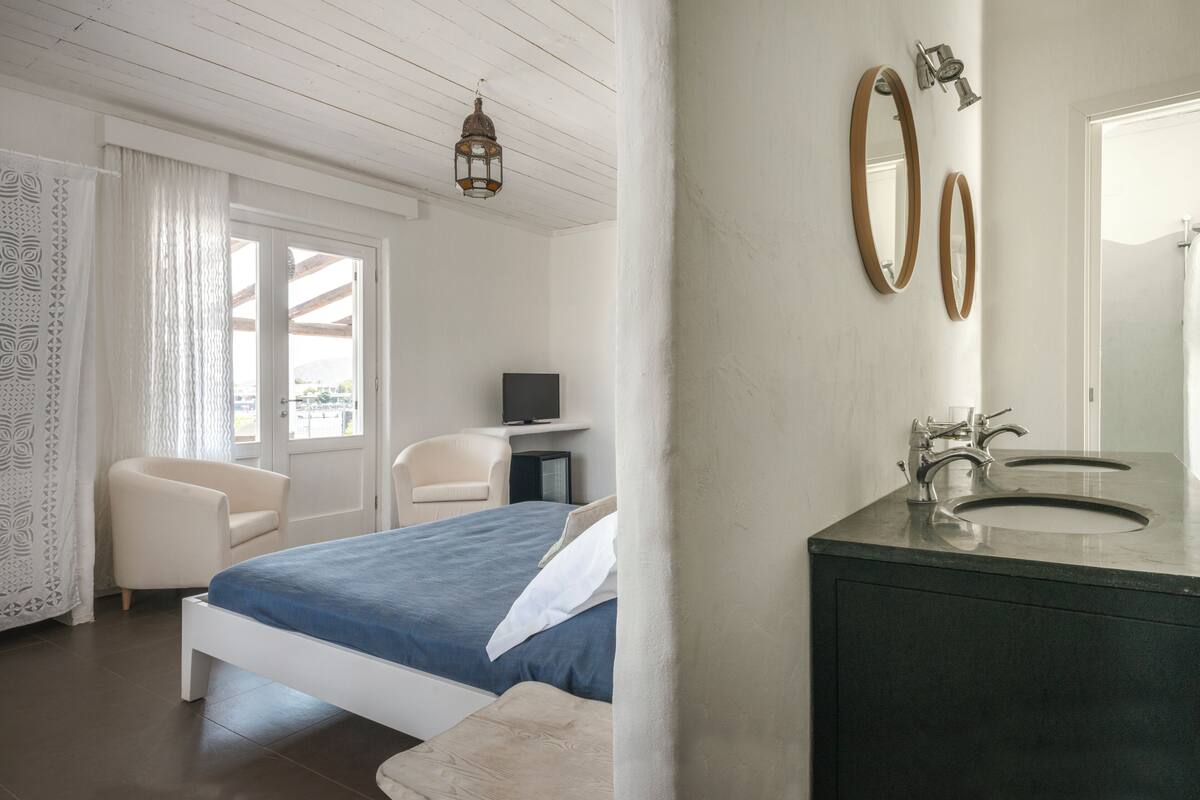 A bedroom with a bed, a chair, a desk with a TV, and a bathroom with a sink and mirror.