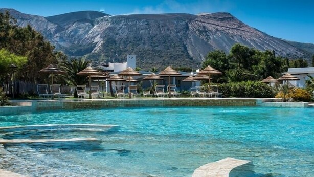 Ein Pool mit klarem Wasser, Liegestühlen, Sonnenschirmen und einem Berg im Hintergrund.
