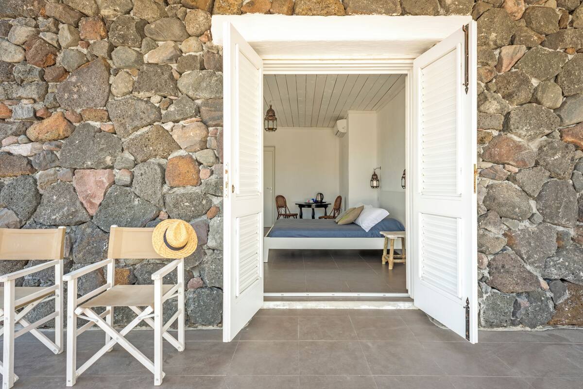 An open doorway leading to a room with a bed, a small table, and two chairs. A straw hat is placed on one of the chairs.