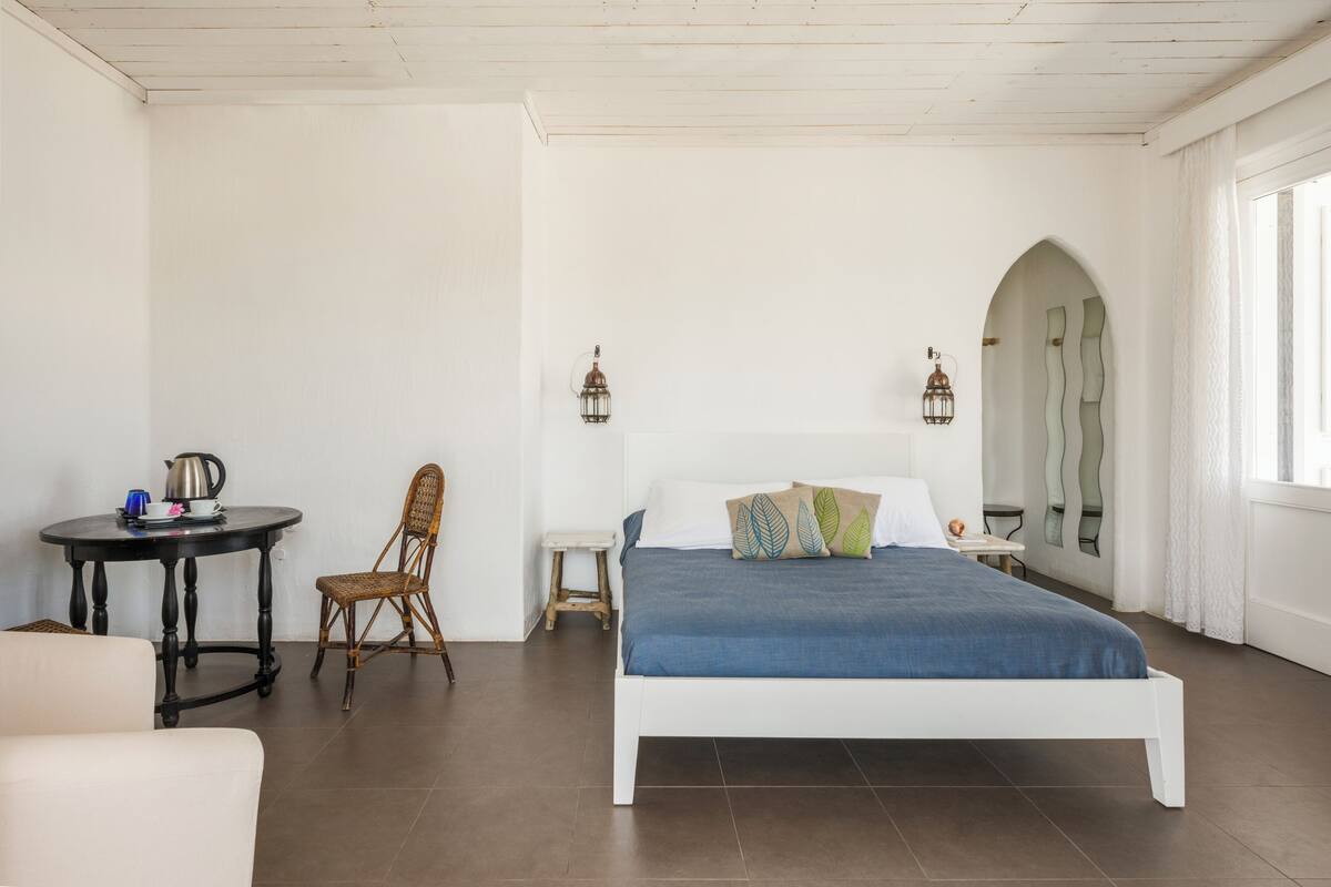 A bedroom with a white bed, a round table with a kettle, a wicker chair, and a small stool.