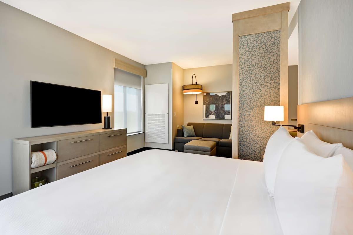 Ein modernes Hotelzimmer mit einem großen Bett, einem Flachbildfernseher, einem Schreibtisch mit Lampe sowie einer Sitzecke mit Sofa und Wandverkleidung mit Muster.