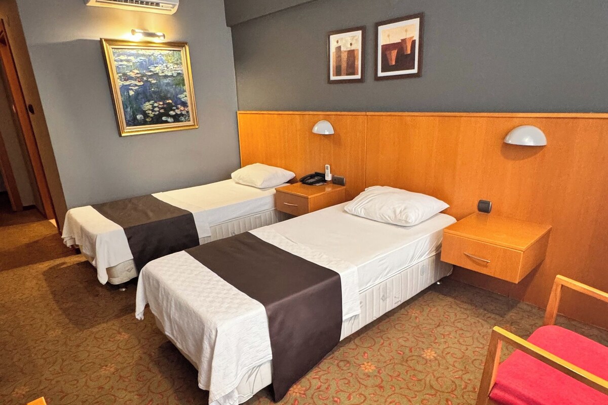 A hotel room with two single beds, a wooden headboard, a nightstand with a lamp, a small table, a chair, and a painting on the wall.