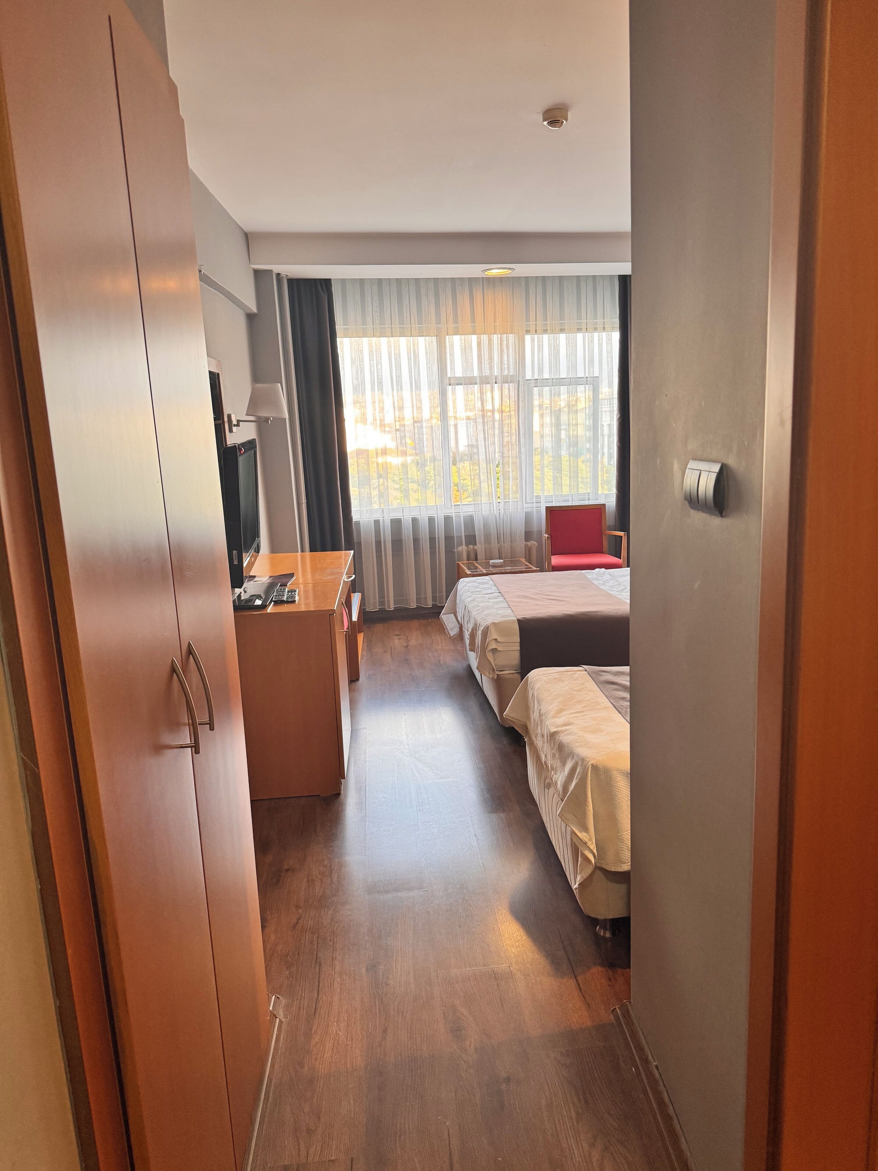 Habitación de hotel con un armario de madera, un escritorio con lámpara, cama y una silla.